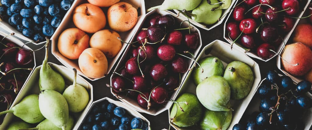 Assortiment de fruits frais d'été en barquettes : cerises, myrtilles, abricots, poires et raisins avec gouttes d'eau.