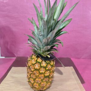 Ananas entier mûr jaune-vert avec sa couronne verte sur fond rose froissé et papier brun. Une étiquette sombre est attachée.