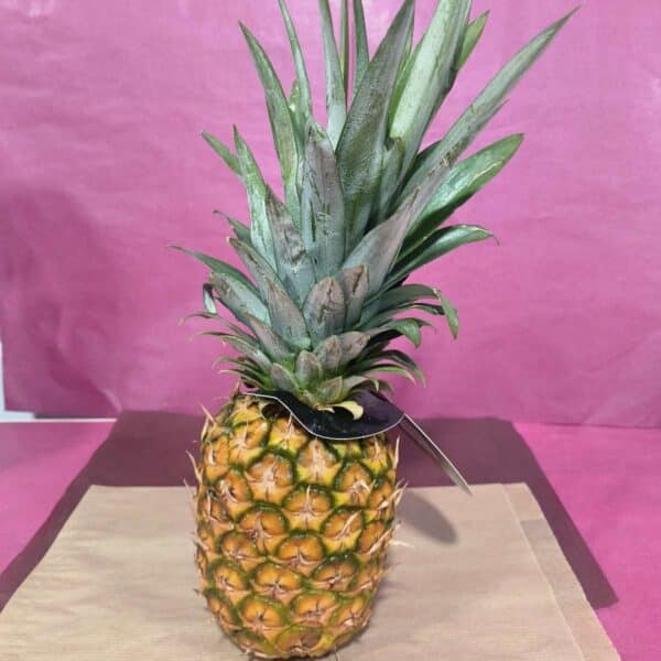 Ananas entier mûr jaune-vert avec sa couronne verte sur fond rose froissé et papier brun. Une étiquette sombre est attachée.