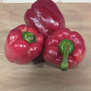 Trois poivrons rouges frais, l'un grand et deux plus petits avec des tiges vertes, posés sur une surface en bois clair.