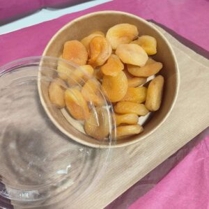 Bol en papier kraft rempli d'abricots secs orange, avec un couvercle transparent marqué '100% PET' sur un fond rose et brun.