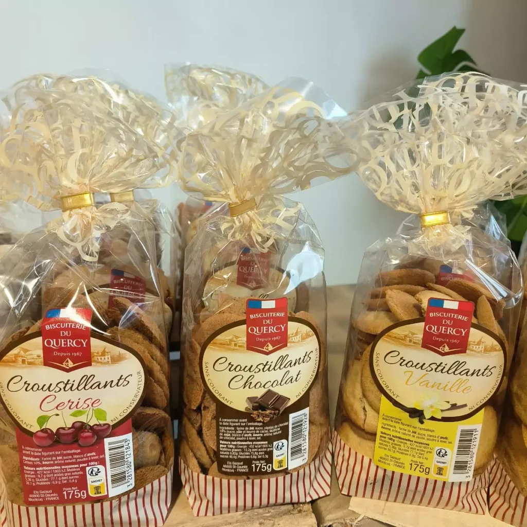 Trois sacs de biscuits Croustillants Biscuiterie du Quercy, saveurs Cerise, Chocolat et Vanille, chacun de 175g. Emballages transparents avec rubans dorés.