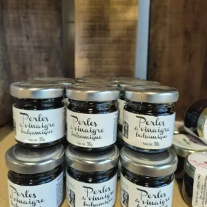 Petits bocaux de Perles de vinaigre balsamique empilés, chacun indiquant "Poids net: 30g". Arrière-plan flou de planches en bois.