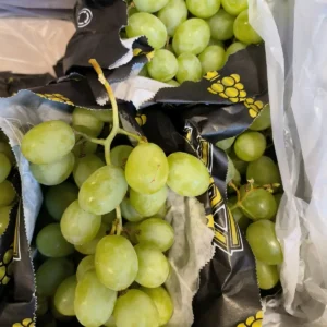 Grappes de raisins verts frais dans un emballage noir orné d'un logo de grappe et d'un symbole de recyclage, près d'un sac plastique blanc.