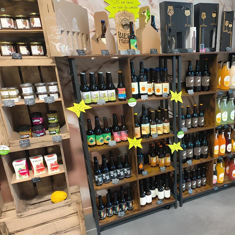 Boutique de produits locaux avec étagères bois et métal. Bouteilles de bières artisanales, jus, confitures et thés PAGES. Texte visible: BIER FRAI.