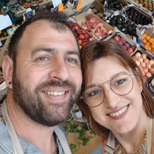 Couple souriant au marché, un homme barbu et une femme aux lunettes posant devant des étals de fruits frais variés.