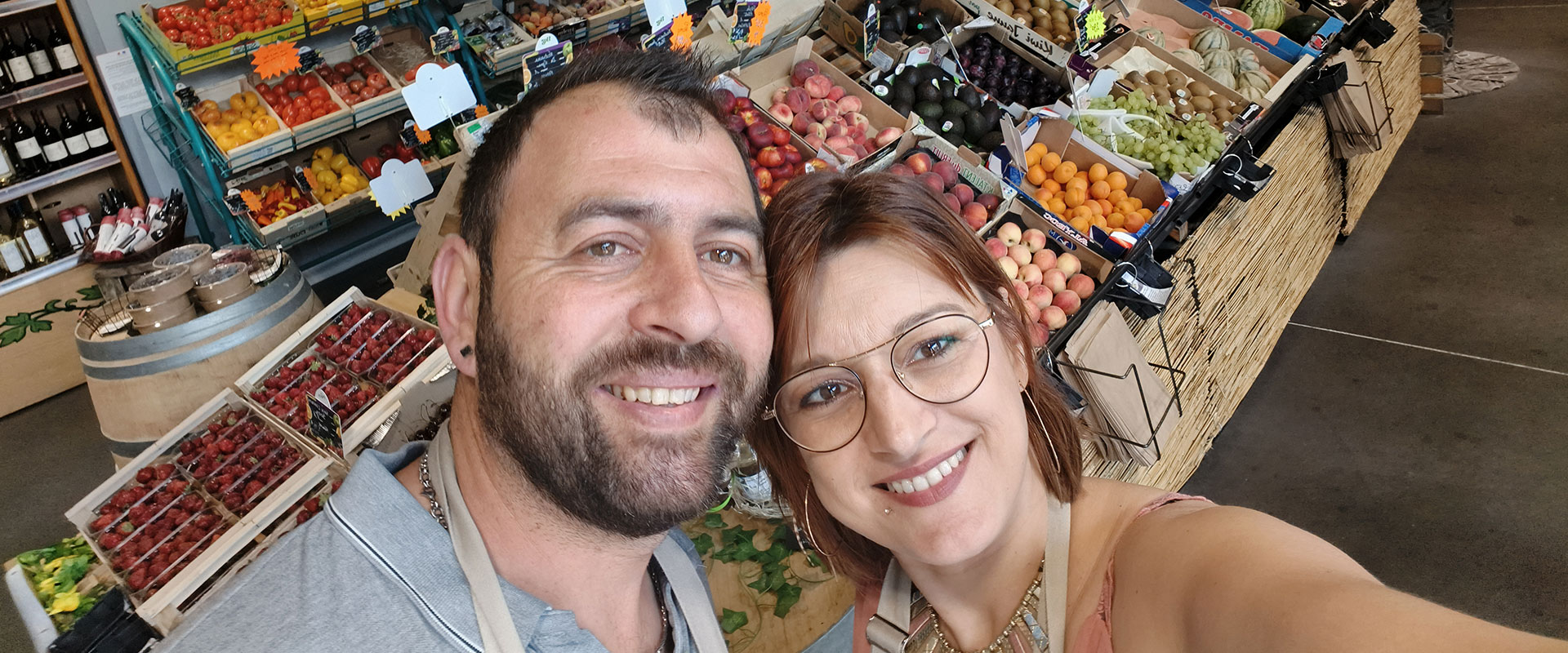 Couple souriant en selfie devant un étal de fruits et légumes frais: fraises, tomates, pêches, avocats, raisins. Rayons de vin visibles.