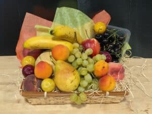 Corbeille en osier garnie d'un assortiment coloré de fruits frais : bananes, raisins, cerises, prunes, abricots, poire, pomme. Sur table en bois.