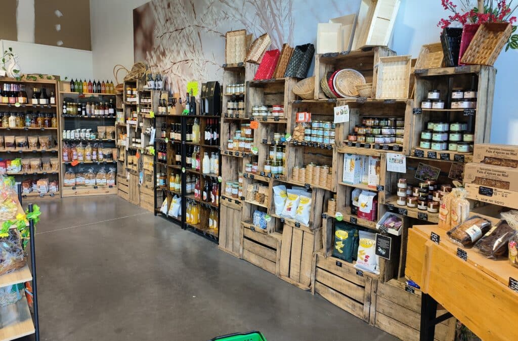 Épicerie avec des étagères et caisses en bois présentant des produits alimentaires variés : bocaux, Pain d'épices au Miel, bouteilles, chips Sauvage, paniers et mural végétal.