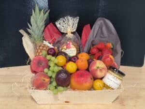 Panier garni de fruits frais (ananas, fraises, pêches, raisins, mangue), biscuits Quercy et miel de Tournesol Apisphère dans une caisse en bois.